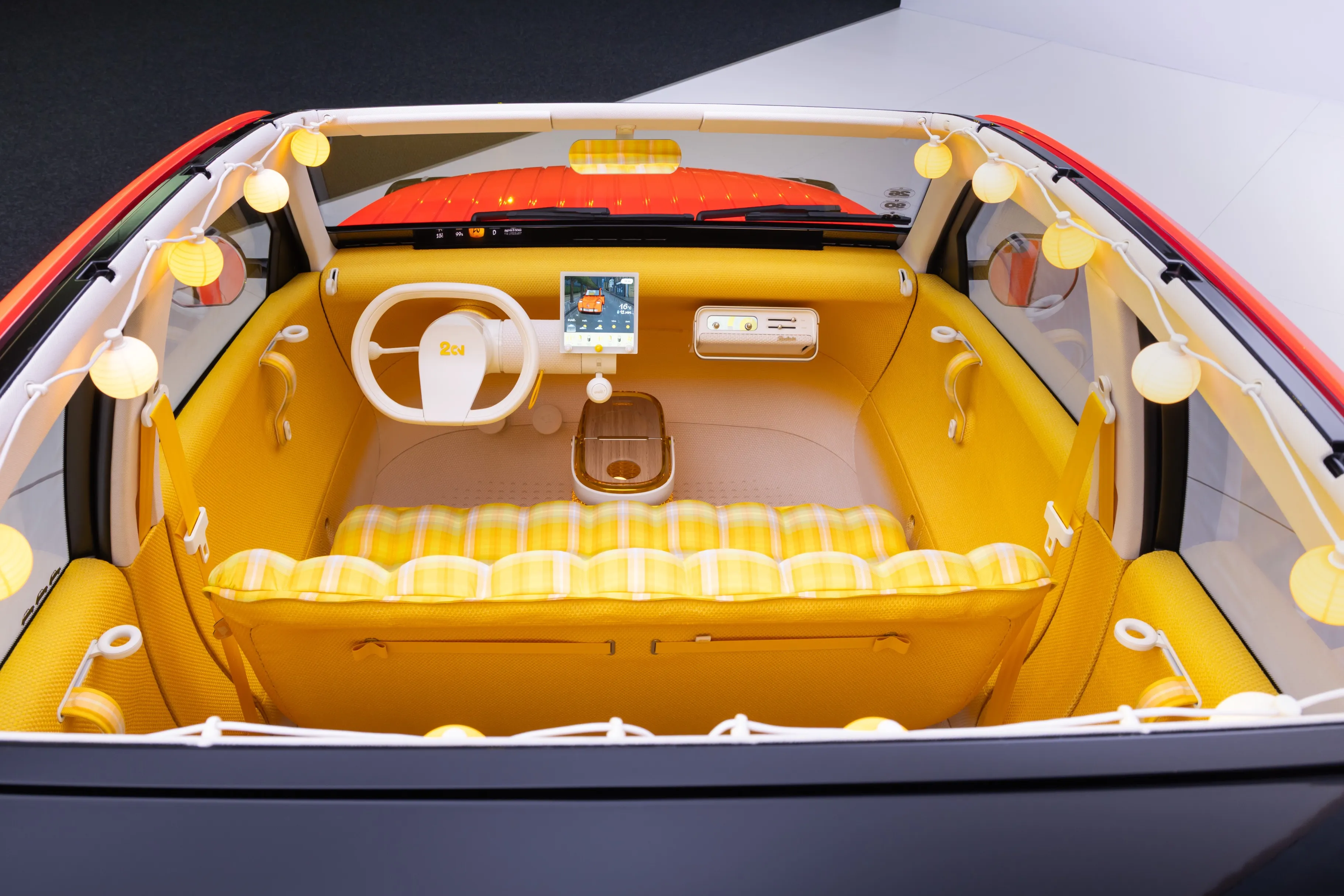 An overhead view of the interior of the Citroën 2CV concept car. Looking in through the open roof, we see a mostly bright yellow interior with a striped bench seat, white pill-shaped steering wheel, and globe lights that hang along the side and rear edges of the roofline.