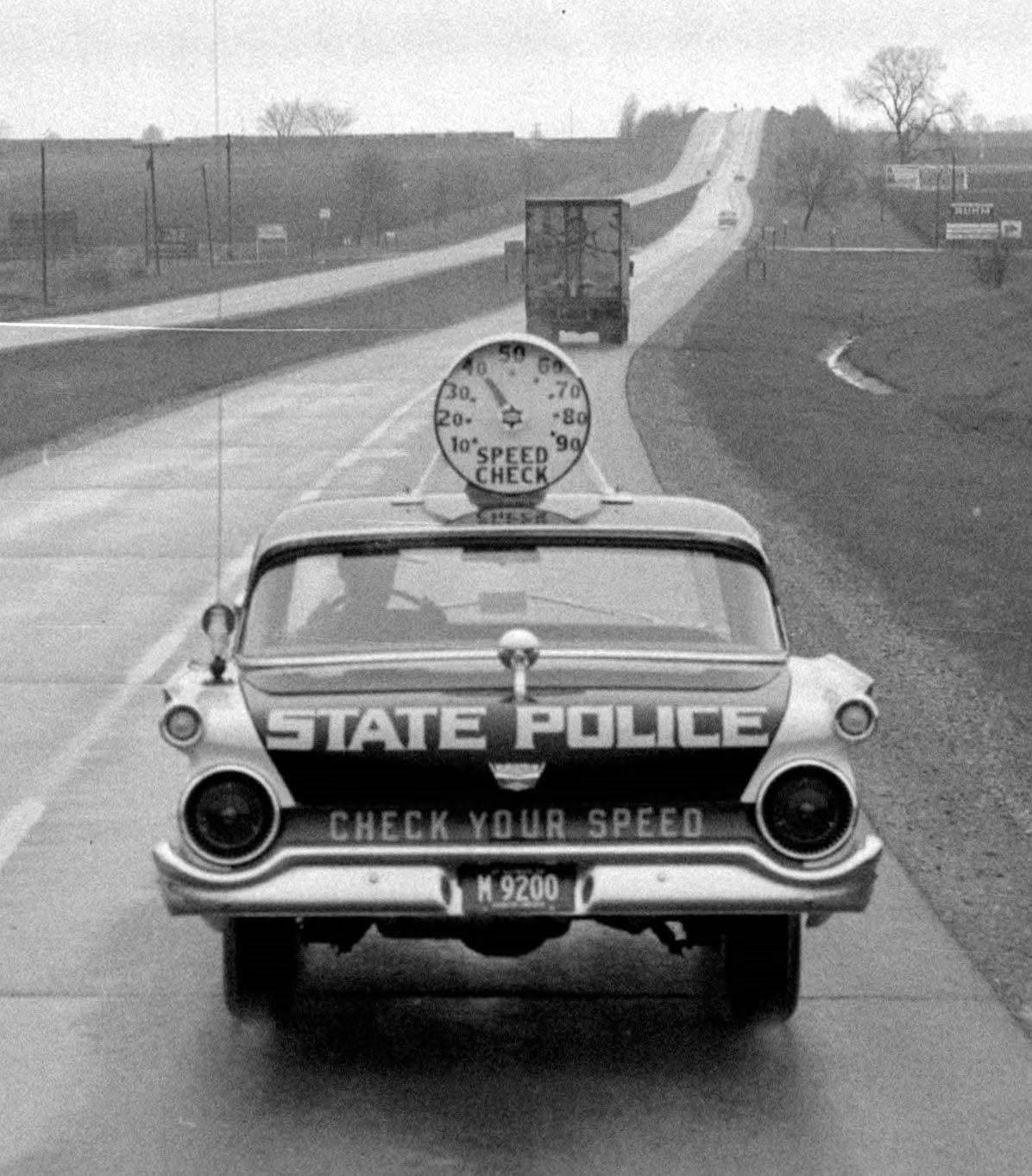 A black and white photo of the rear of an old (1950s–1960s) state police car driving on a road. A large round analog spedometer is mounted on the roof, facing backwards, with the words 'speed check' written below the dial.  On the back of the car it says 'state police' in large letters with 'check your speed' written below in smaller letters.